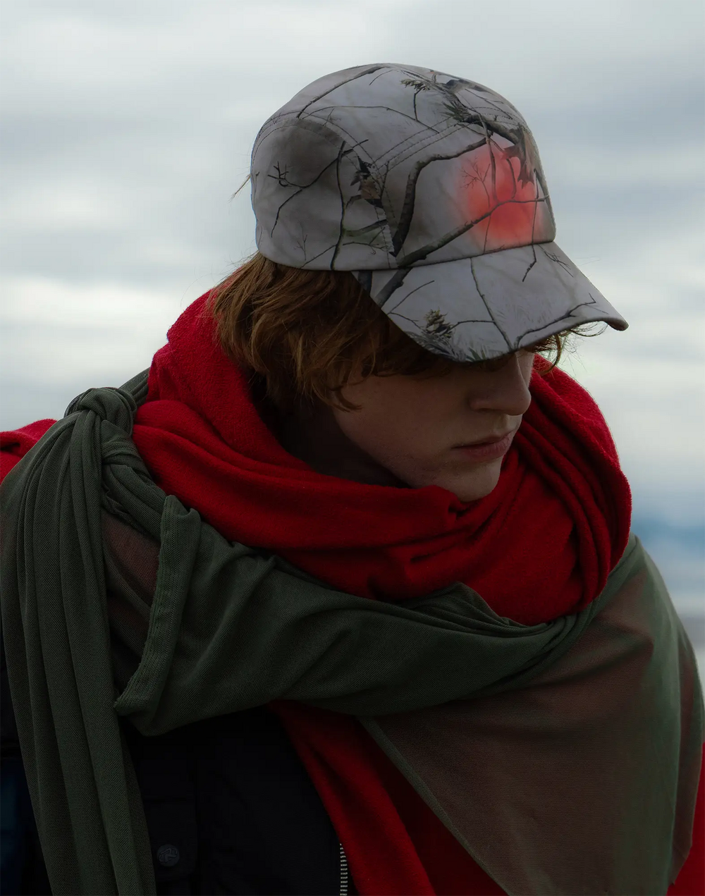 Person wearing a camouflage cap and red scarf against a cloudy sky